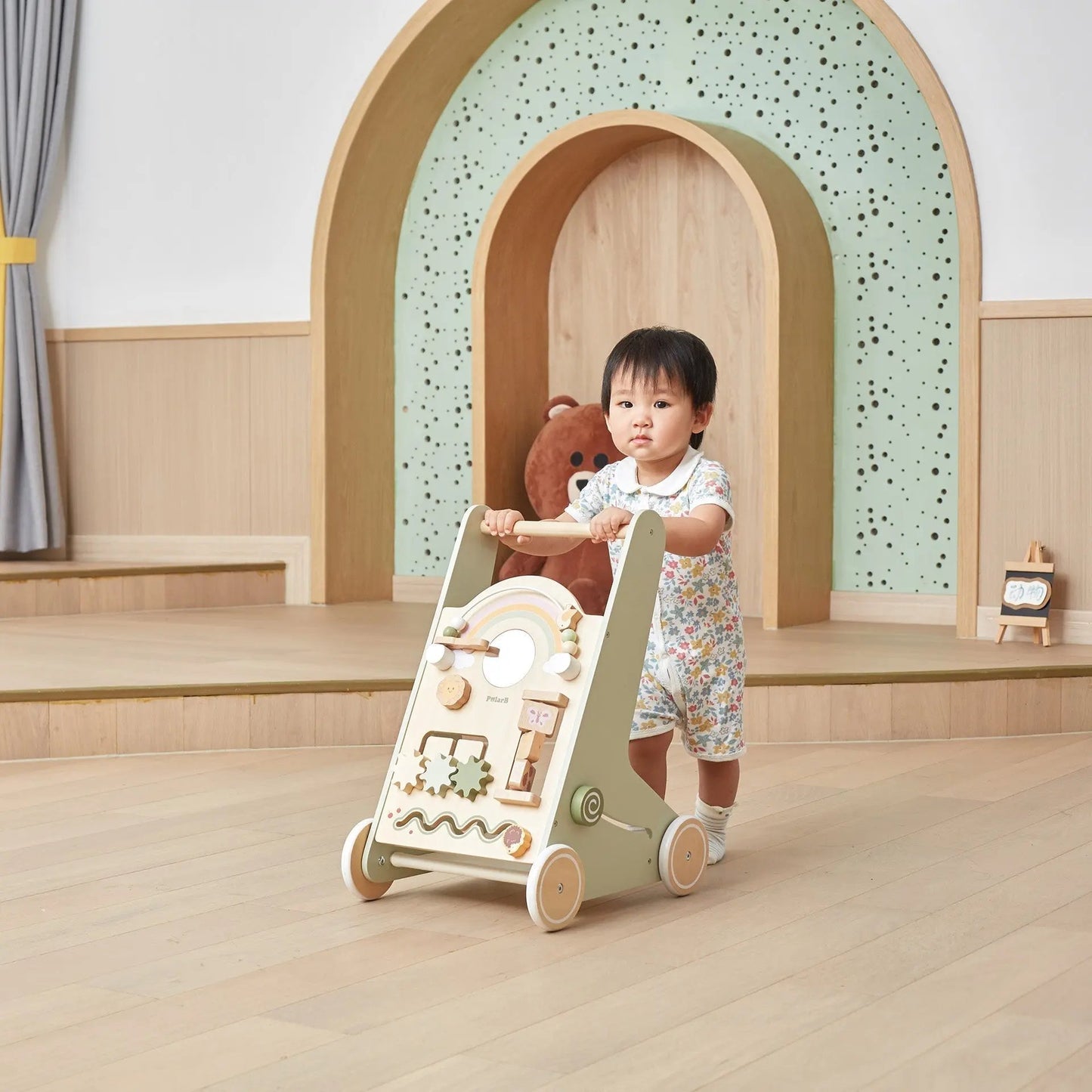 Child playing with a wooden toy walker in a room with a teddy bear and decorative arches.