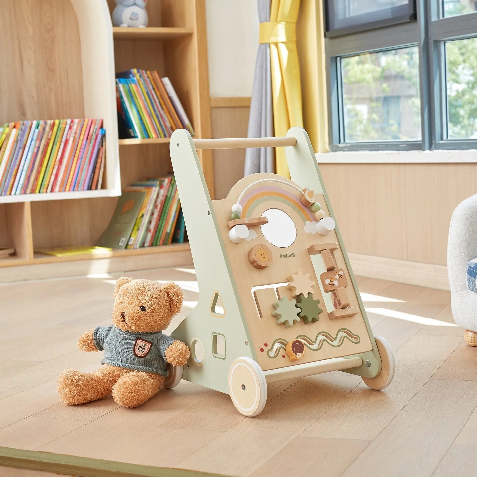 Wooden toy push cart with educational panels and a teddy bear on a wooden floor in a room with books and a window.