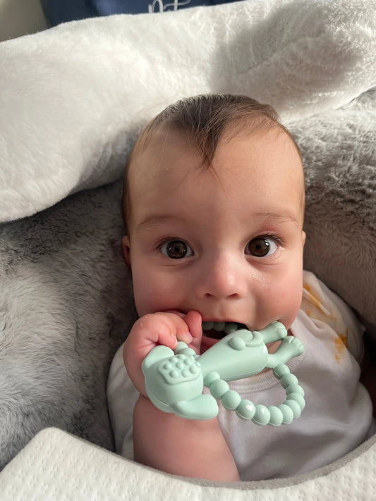 Baby holding a blue teething toy with a soft white blanket in the background
