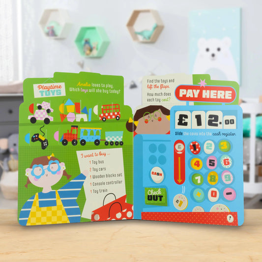A children's interactive cardboard toy money playset displayed open on a light wooden tabletop, featuring colourful printed pages with toy icons, a cash register design, and cartoon characters in a softly blurred room background.