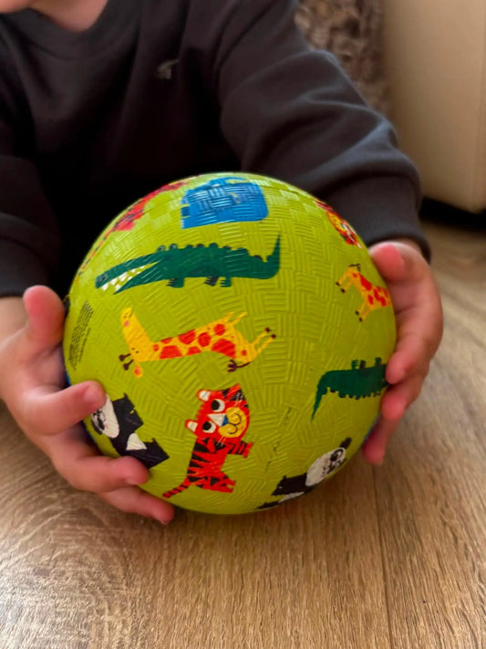 A child's hands holding a lime green textured ball decorated with colourful cartoon animal illustrations, set against a home background with a wooden floor.