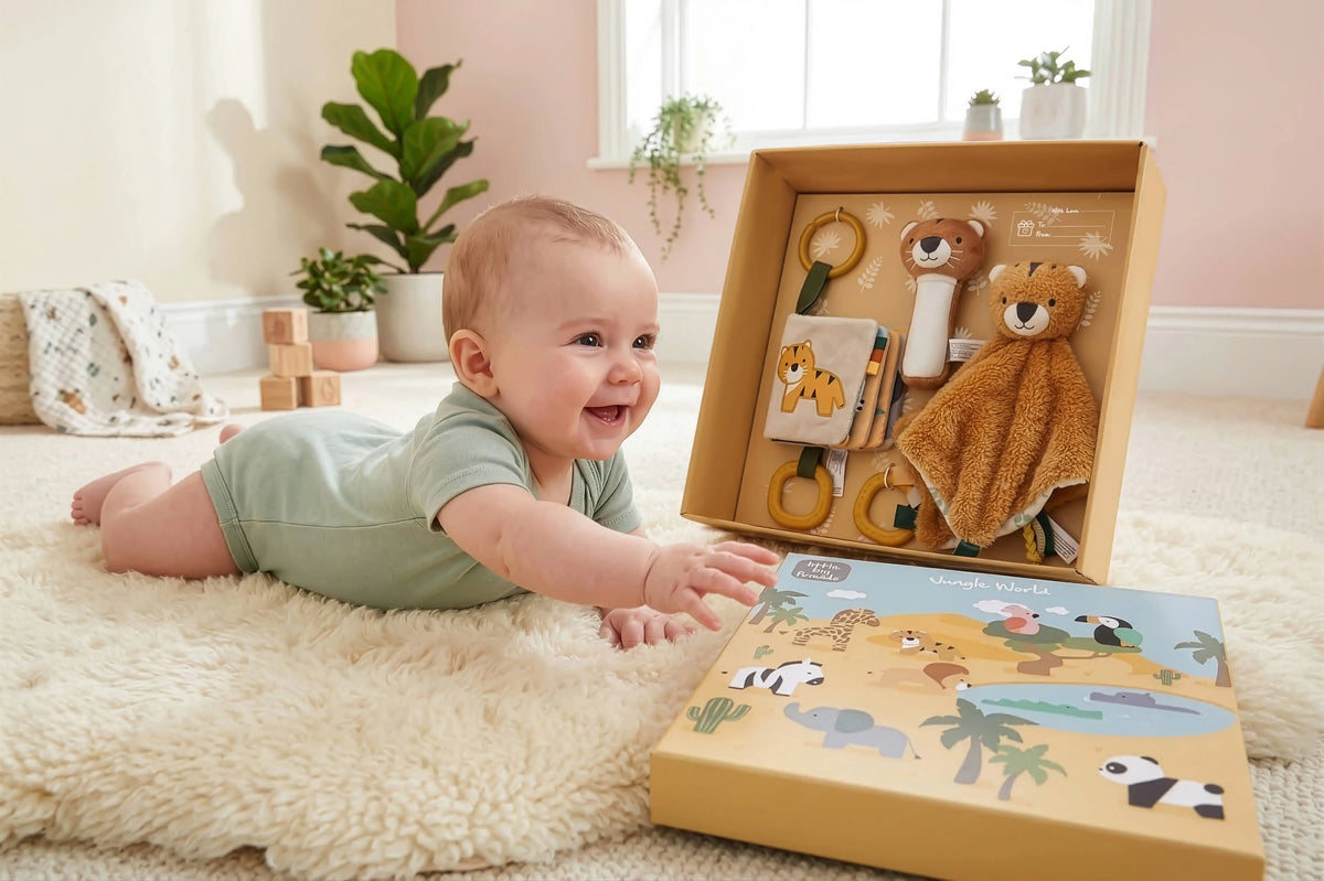 A baby in a light green outfit lies on a cream rug, smiling at an open safari-themed sensory toy gift box. The box contains a plush teddy bear comforter, a giraffe teething ring, and other toys, with a matching illustrated gift box in front.