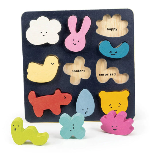 A wooden children's puzzle board with colorful cutout shapes and words printed on a white background. The board is dark blue with cutouts for a white cloud, pink bunny, tan cloud labeled 'happy', blue sad face, yellow chick, tan flower labeled 'content', tan cloud labeled 'surprised', red dog, light blue teardrop, yellow bear, and three loose matching pieces in green, light blue, and pink.
