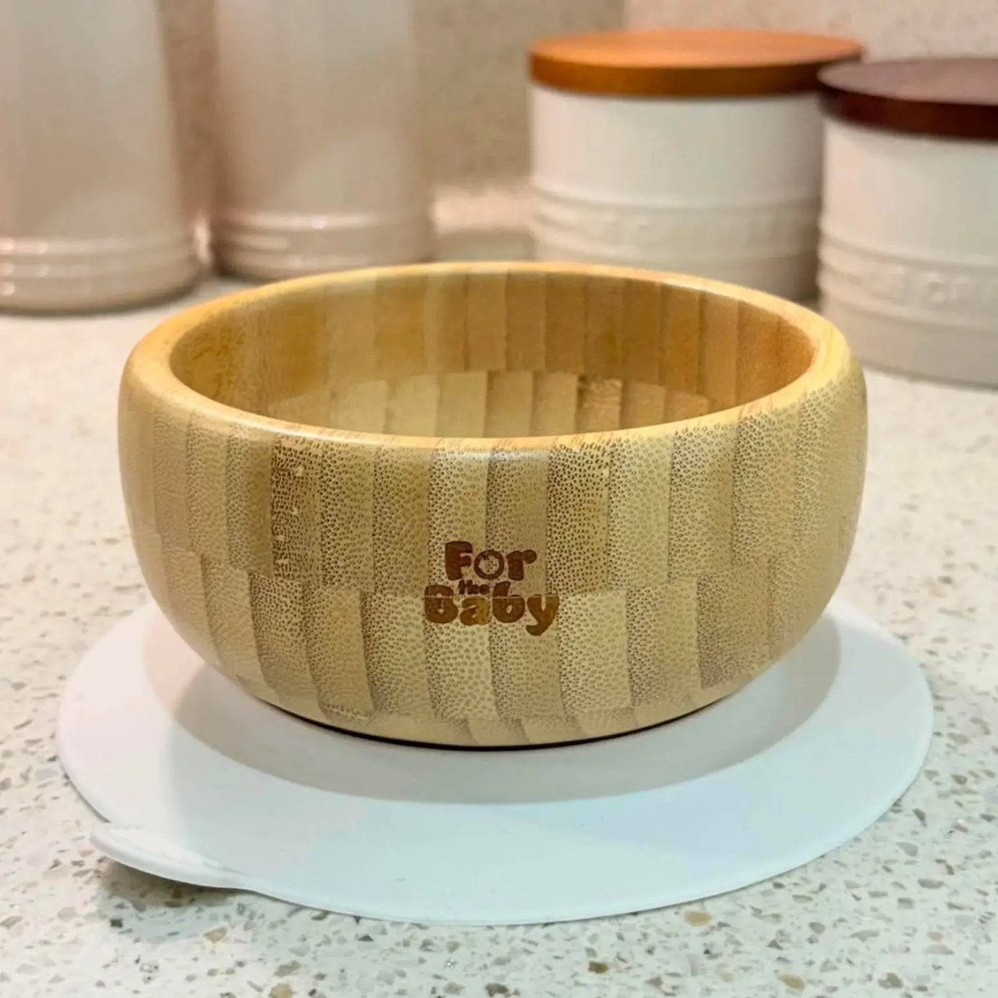 A light natural bamboo baby bowl with a textured basketweave pattern and the words 'For the Baby' engraved on the front, resting on a white plate on a speckled countertop.
