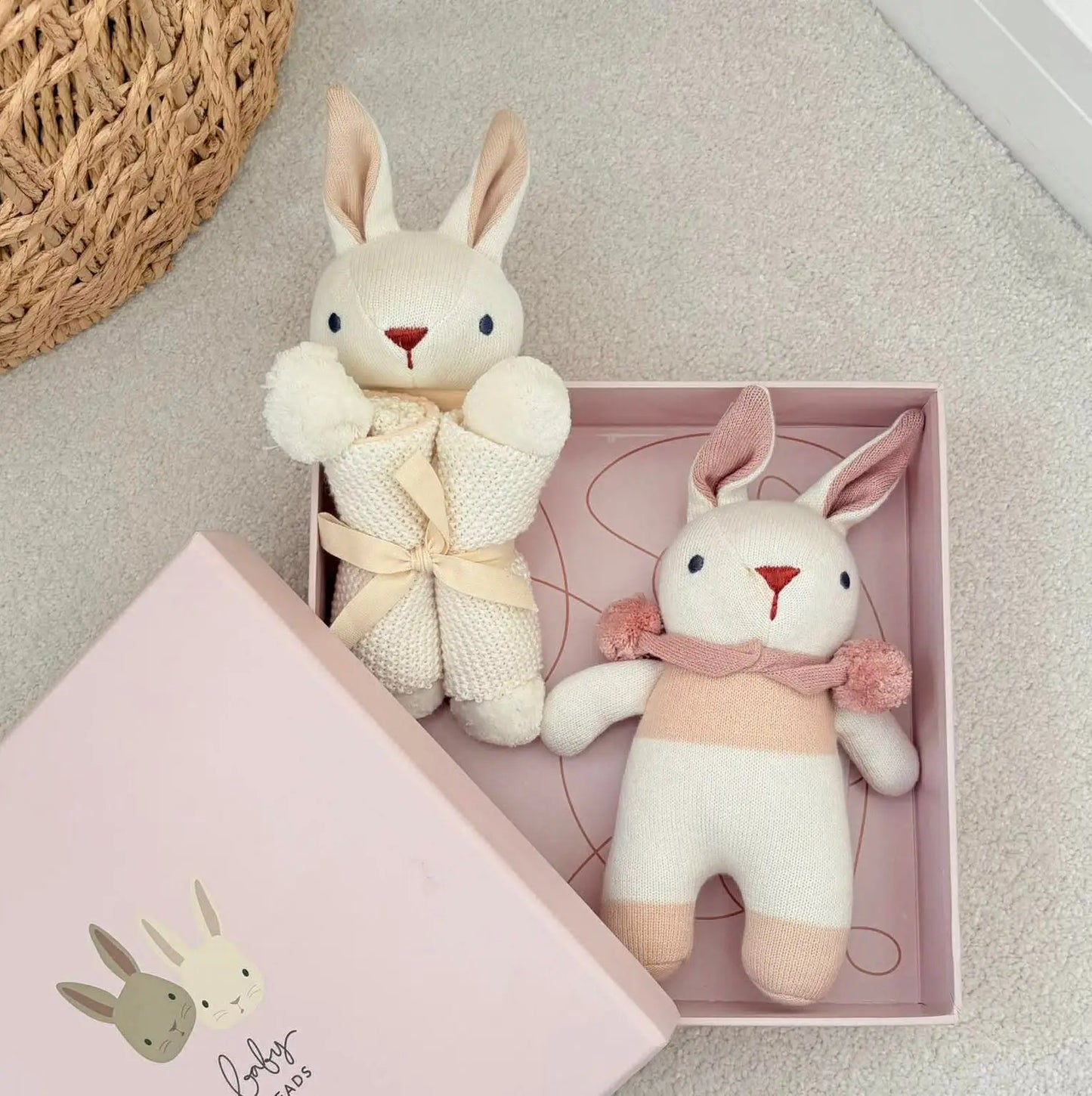 Two plush rabbit toys in a pink box on a textured surface