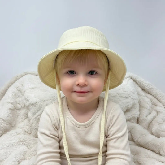 Child wearing a lemon coloured hat sitting on a soft surface with a plain background