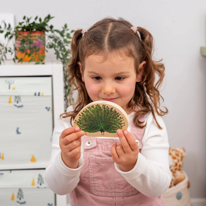 Young girl holding a wooden block with a peacock design in a room with plants and furniture.