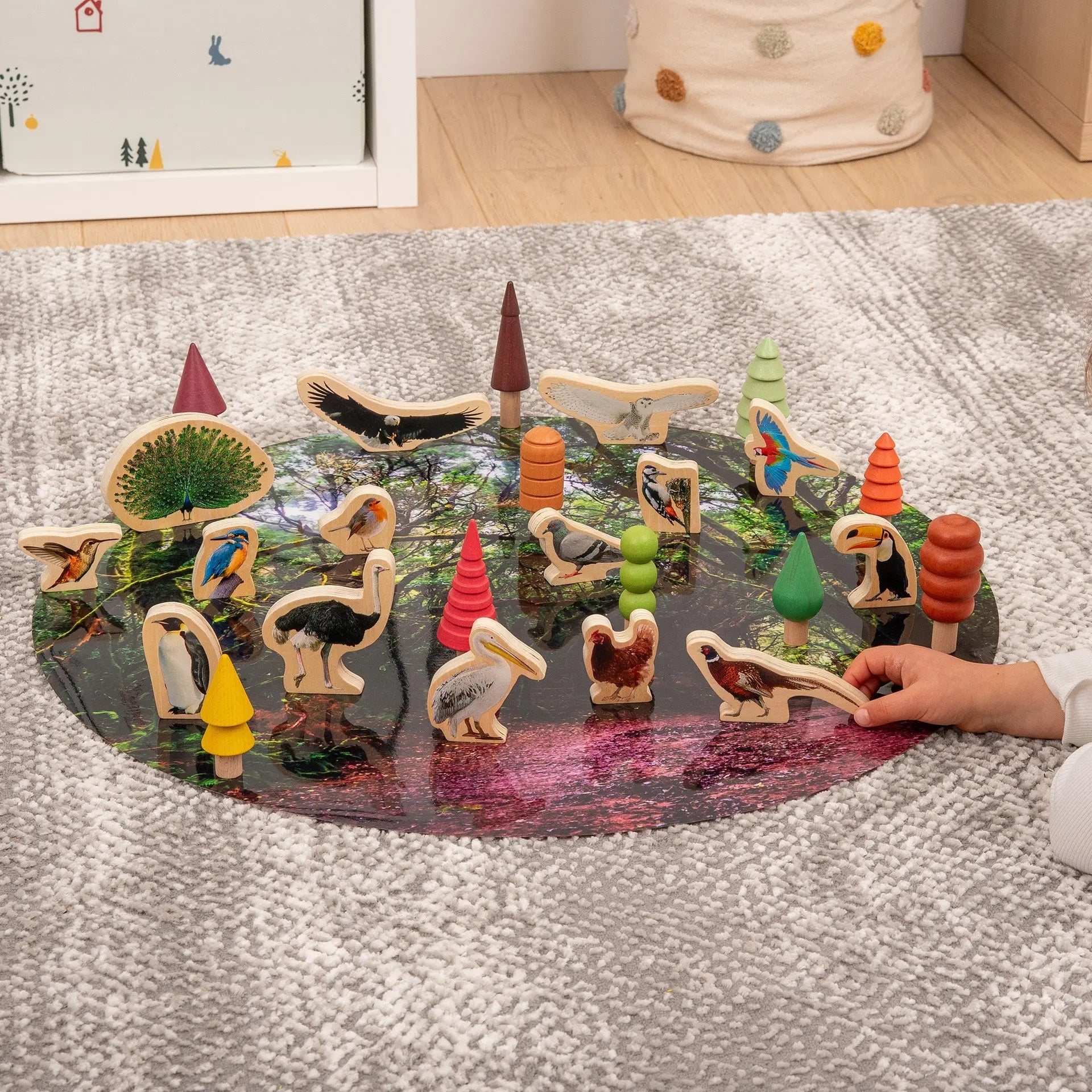 Children's wooden animal toys on a round mat with a hand interacting with one of them, set against a neutral background.