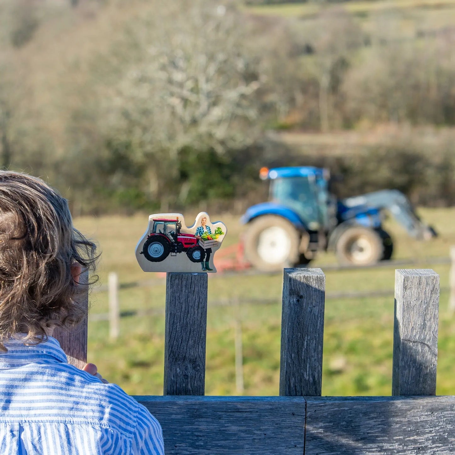 Person looking at a toy figure of a woman on a tractor with a real tractor in the background.