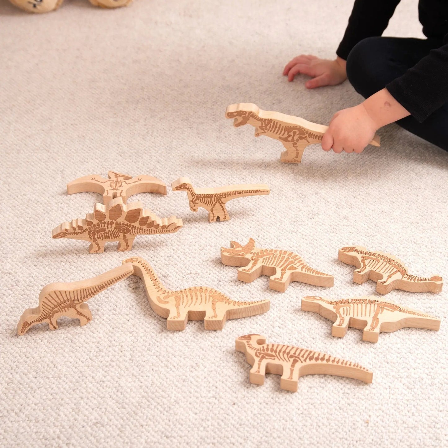 Wooden dinosaur fossils on a carpeted floor with a child's hand reaching for one.