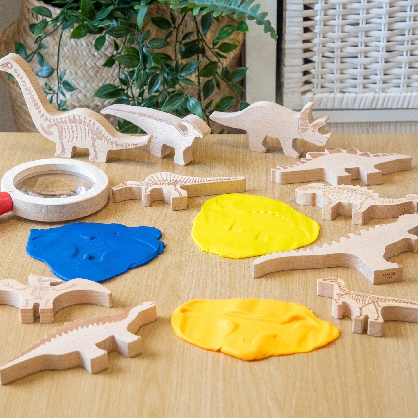 Wooden dinosaur toys and playdough on a wooden table with a plant in the background.