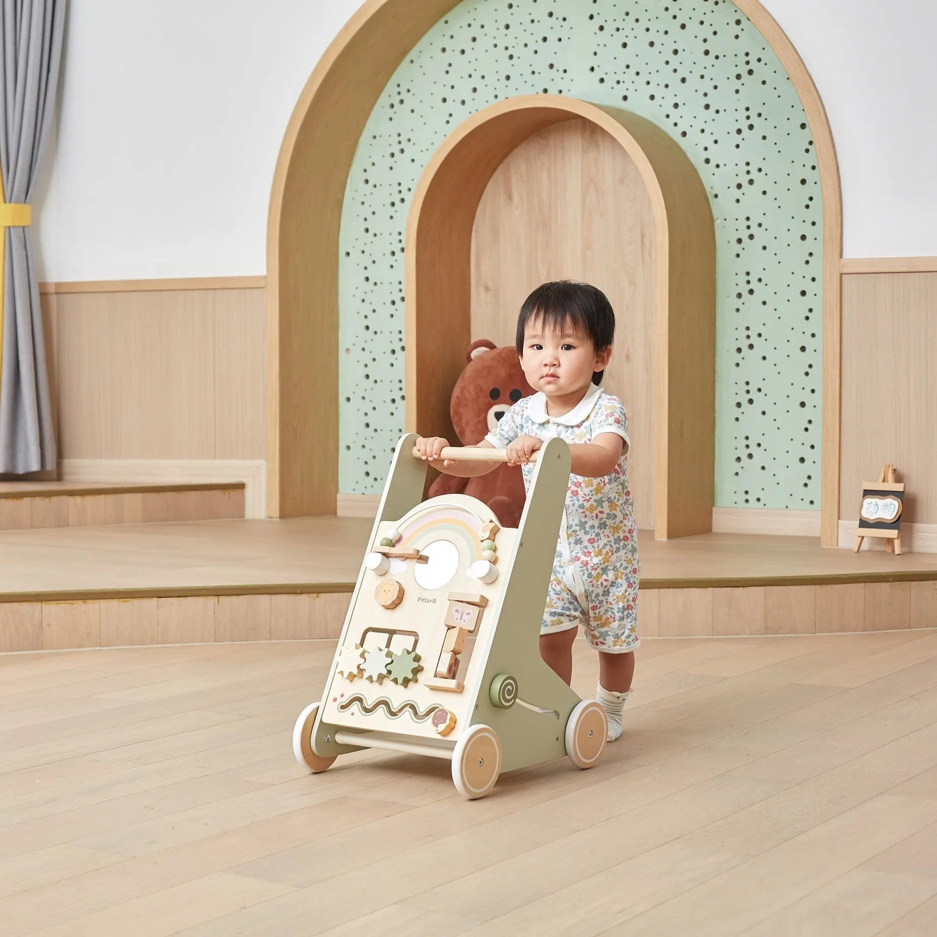 Child playing with a wooden toy walker in a room with a teddy bear and decorative arches.