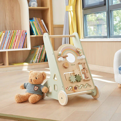 Wooden toy push cart with educational panels and a teddy bear on a wooden floor in a room with books and a window.