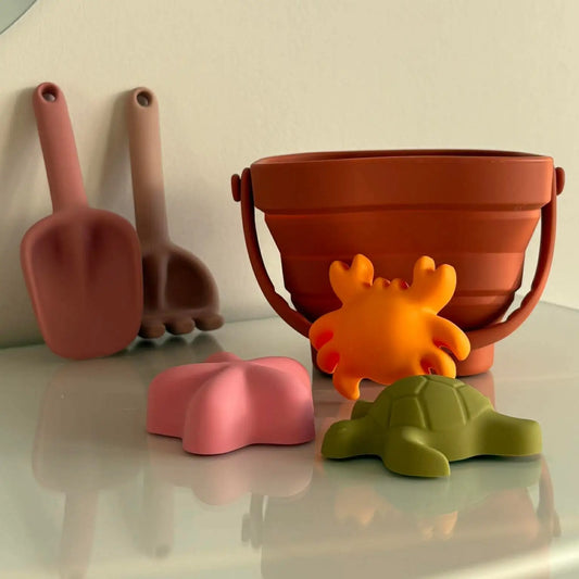 Set of colourful children's sand toys including a bucket, shovels, and sea creature moulds on a reflective surface.