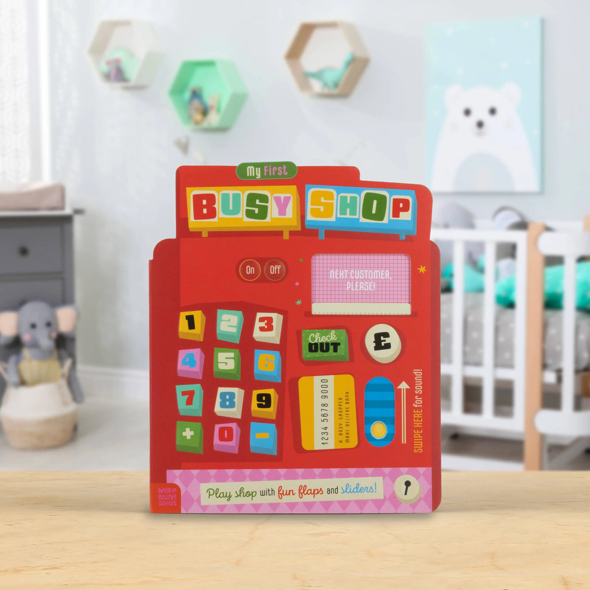A children's toy 'My first BUSY SHOP' wooden activity board placed on a light wood table in a softly blurred nursery setting. The red toy features colourful buttons, a cash register display, a keypad, and shopping-themed graphics.