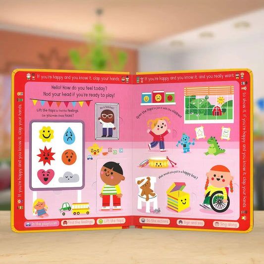 A children's educational picture book with bright pink and red covers, displayed open on a light wooden surface with a softly blurred indoor background. The pages are filled with colourful cartoon illustrations, including faces, animals, vehicles, and simple scenes.