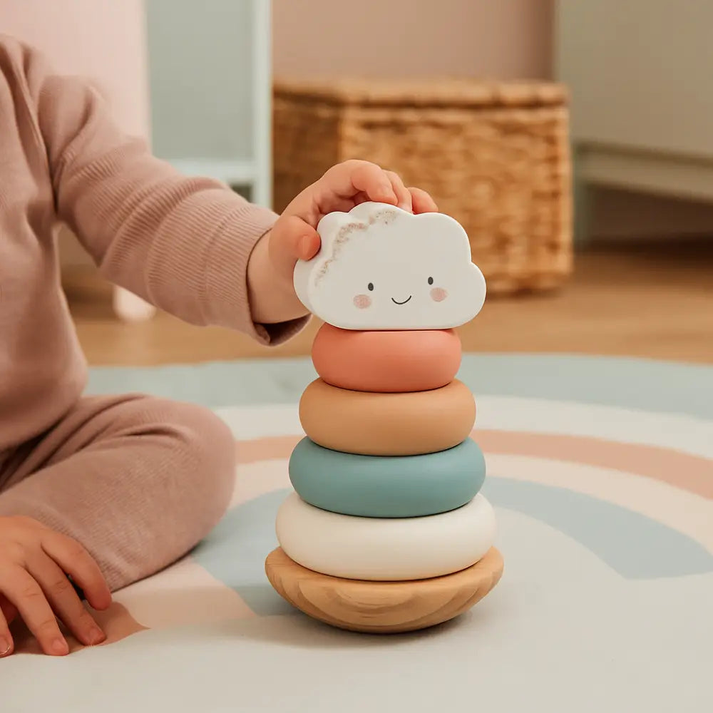 A child in a pink outfit is placing a smiling white cloud-shaped ring on top of a pastel stacking toy set on a play rug in a softly lit room.