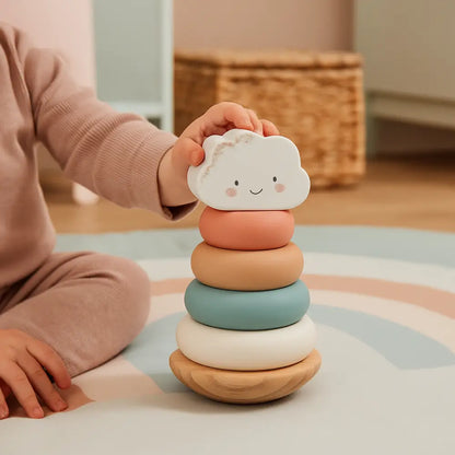 A child in a pink outfit is placing a smiling white cloud-shaped ring on top of a pastel stacking toy set on a play rug in a softly lit room.