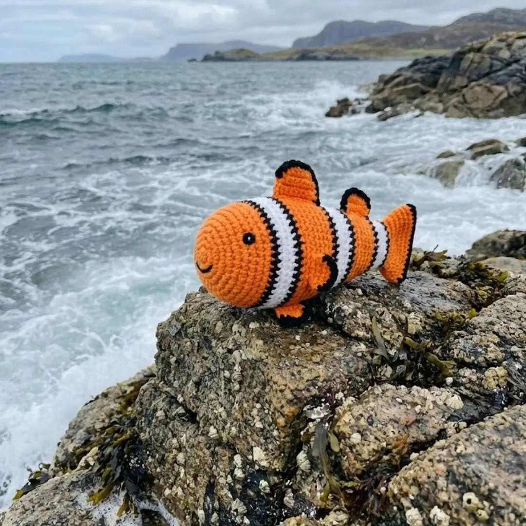 Crocheted clownfish toy on a rock with ocean and mountains in the background