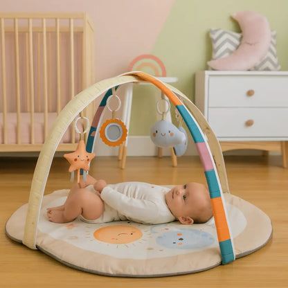 A baby lies on a round play mat under a fabric activity gym in a softly styled nursery. The mat and arch have a cream base with orange, teal, and pink accents, and include sun and cloud graphics.