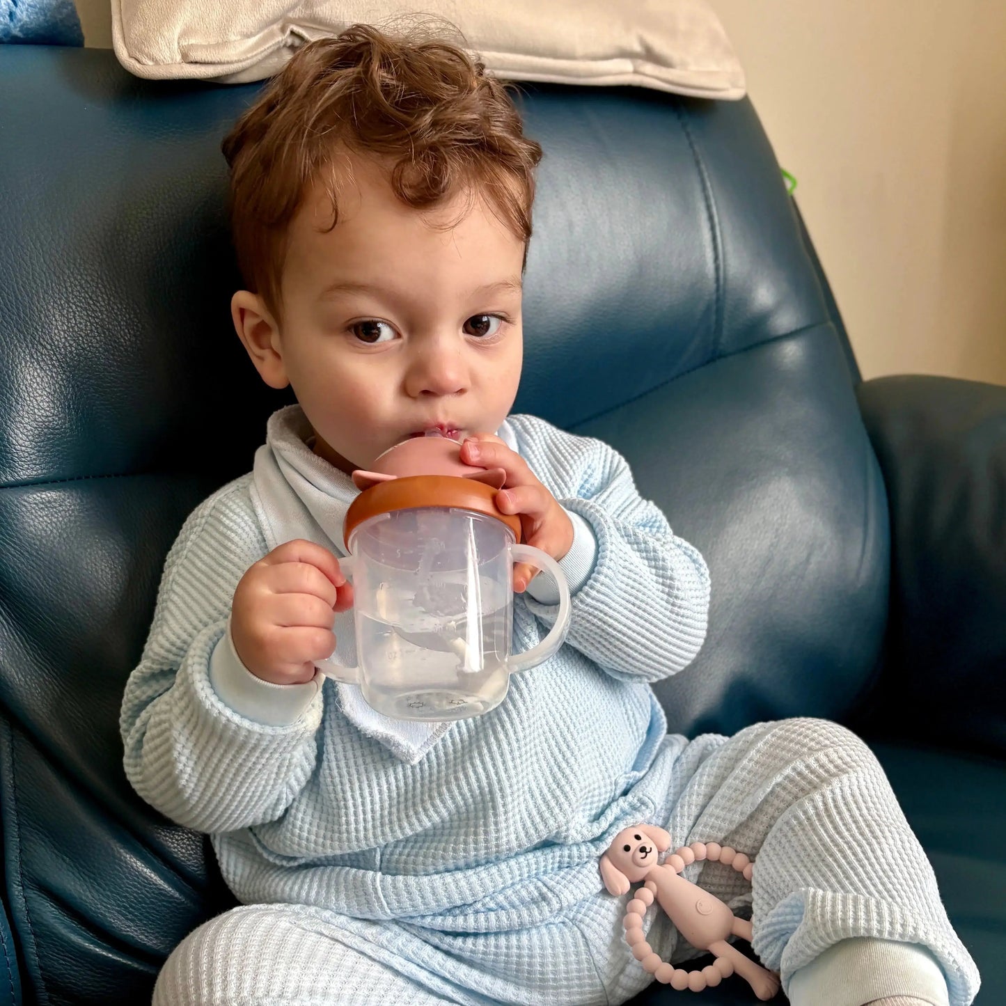 Child in a light blue outfit holding a clear cup with a brown lid on a blue chair.