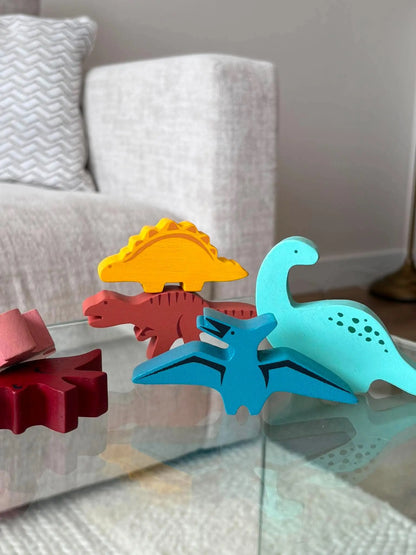 A set of colourful wooden dinosaur toys arranged on a glass table in a living room setting, with a grey sofa and patterned pillow in the background.