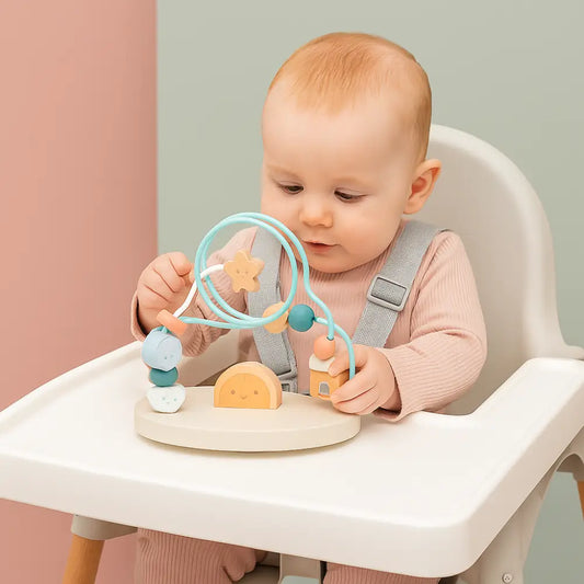 A baby sitting in a white high chair, playing with a wooden bead activity toy on the tray. The toy features light blue loops, wooden beads, and small wooden pieces, set against a soft pink and sage green background.
