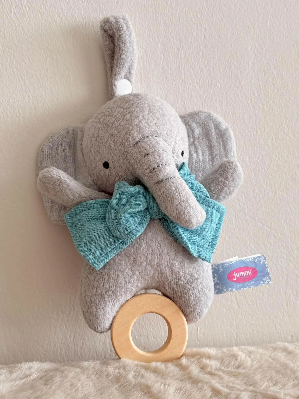 A plush elephant baby toy with a light grey felt body, large floppy ears, a turquoise fabric bow, and a natural wood ring at the bottom, photographed against a plain beige wall.