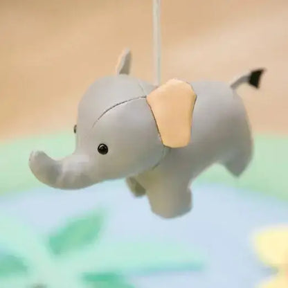 A small plush elephant toy hanging from a white string, with a light grey body, tan ear, and small black tail tip, shown against a softly blurred pastel background.