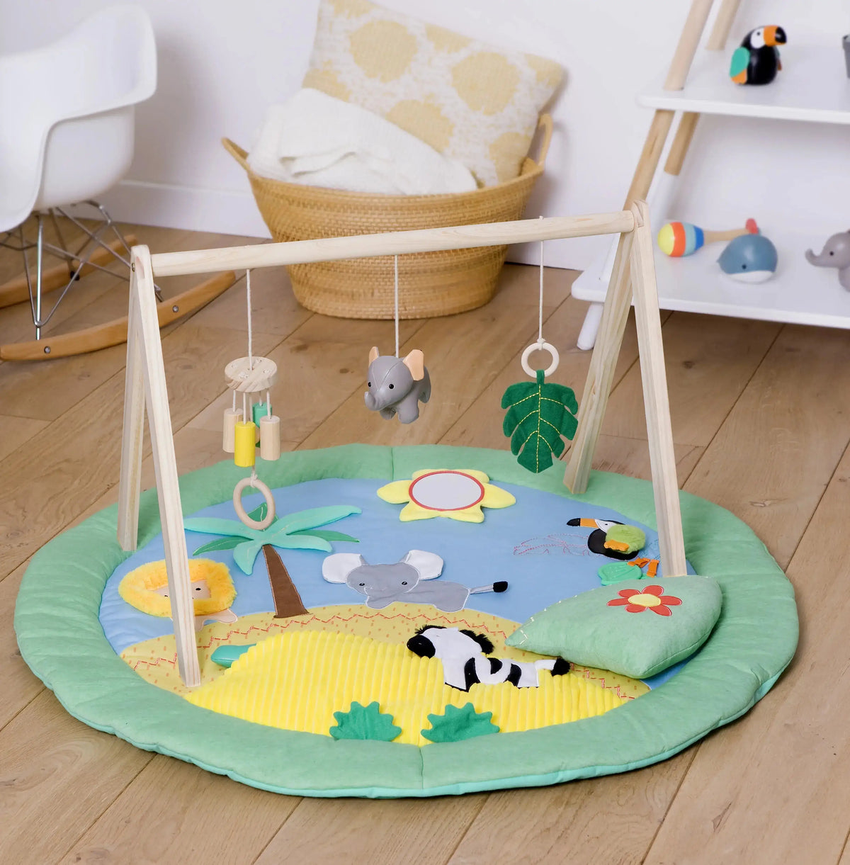 A round baby play mat with a wooden activity gym in a bright nursery setting. The play mat features a safari-themed design with a blue sky, yellow sun, green grass, and animal illustrations including an elephant, lion, zebra, and toucan.