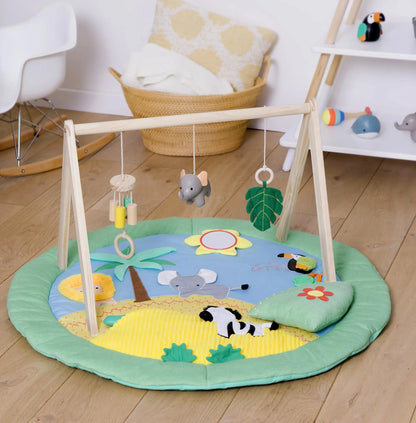 A round baby play mat with a wooden activity gym in a bright nursery setting. The play mat features a safari-themed design with a blue sky, yellow sun, green grass, and animal illustrations including an elephant, lion, zebra, and toucan.