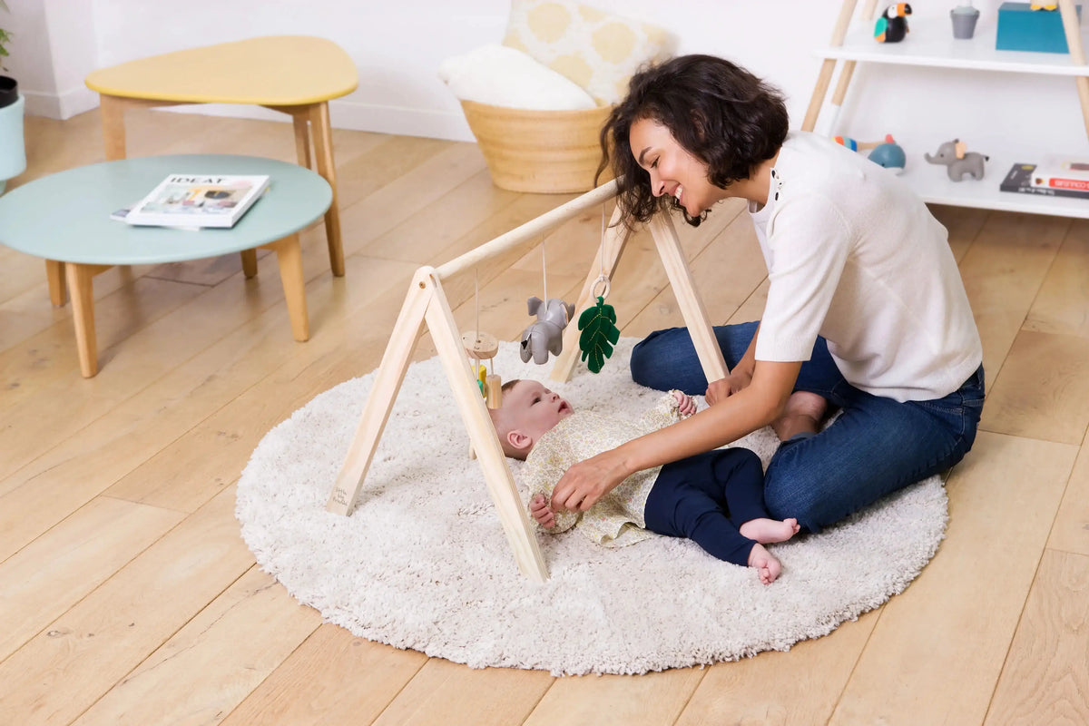 A baby tummy activity gym on a round white shag rug, with a woman kneeling beside it and a baby lying on the floor. The activity gym has a light wood A-frame structure with hanging toys including a green leaf and a grey animal.
