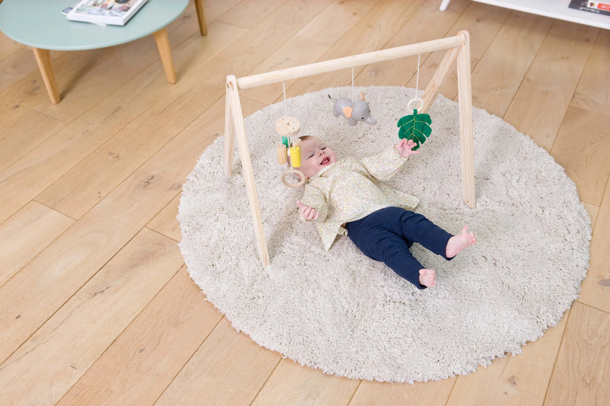 Baby lying on a round rug with a play gym in a room with wooden flooring.