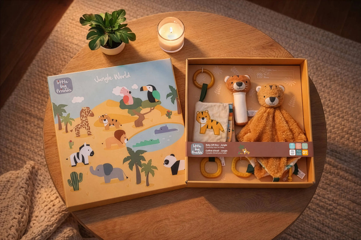 A styled flat-lay of a children's jungle-themed gift set on a round wooden table. The set includes a colourful illustrated book titled 'Jungle World' and an open wooden box containing a plush lion toy, a fabric cloth, a rattle, and a teething ring.