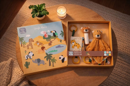 A styled flat-lay of a children's jungle-themed gift set on a round wooden table. The set includes a colourful illustrated book titled 'Jungle World' and an open wooden box containing a plush lion toy, a fabric cloth, a rattle, and a teething ring.