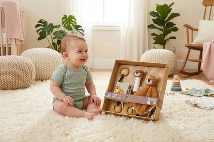 A baby sits on a fluffy cream rug in a bright nursery, smiling next to an open wooden toy gift box containing a plush bear, rings, and other baby toys.