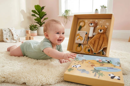 A baby in a light green outfit lies on a cream rug, smiling at an open safari-themed sensory toy gift box. The box contains a plush teddy bear comforter, a giraffe teething ring, and other toys, with a matching illustrated gift box in front.