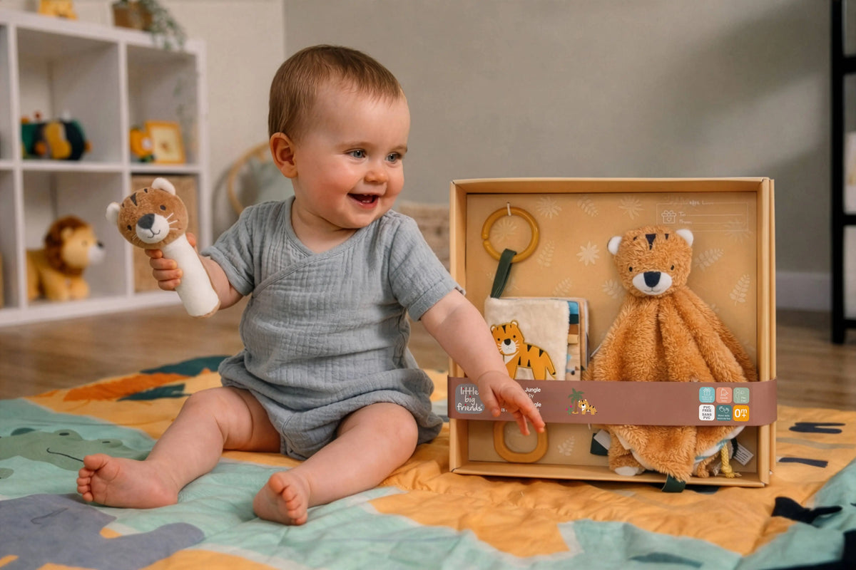 A baby sitting on a colourful play mat holds a plush lion toy while looking at a wooden gift box containing a teddy bear security blanket, a book, and a ring toy. The baby wears a gray waffle-knit romper in a bright nursery setting.