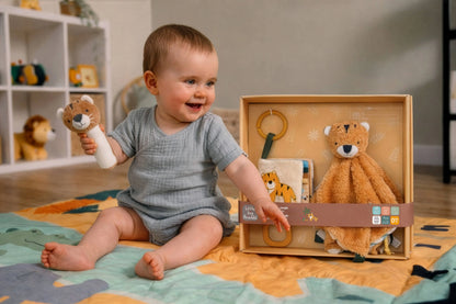A baby sitting on a colourful play mat holds a plush lion toy while looking at a wooden gift box containing a teddy bear security blanket, a book, and a ring toy. The baby wears a gray waffle-knit romper in a bright nursery setting.