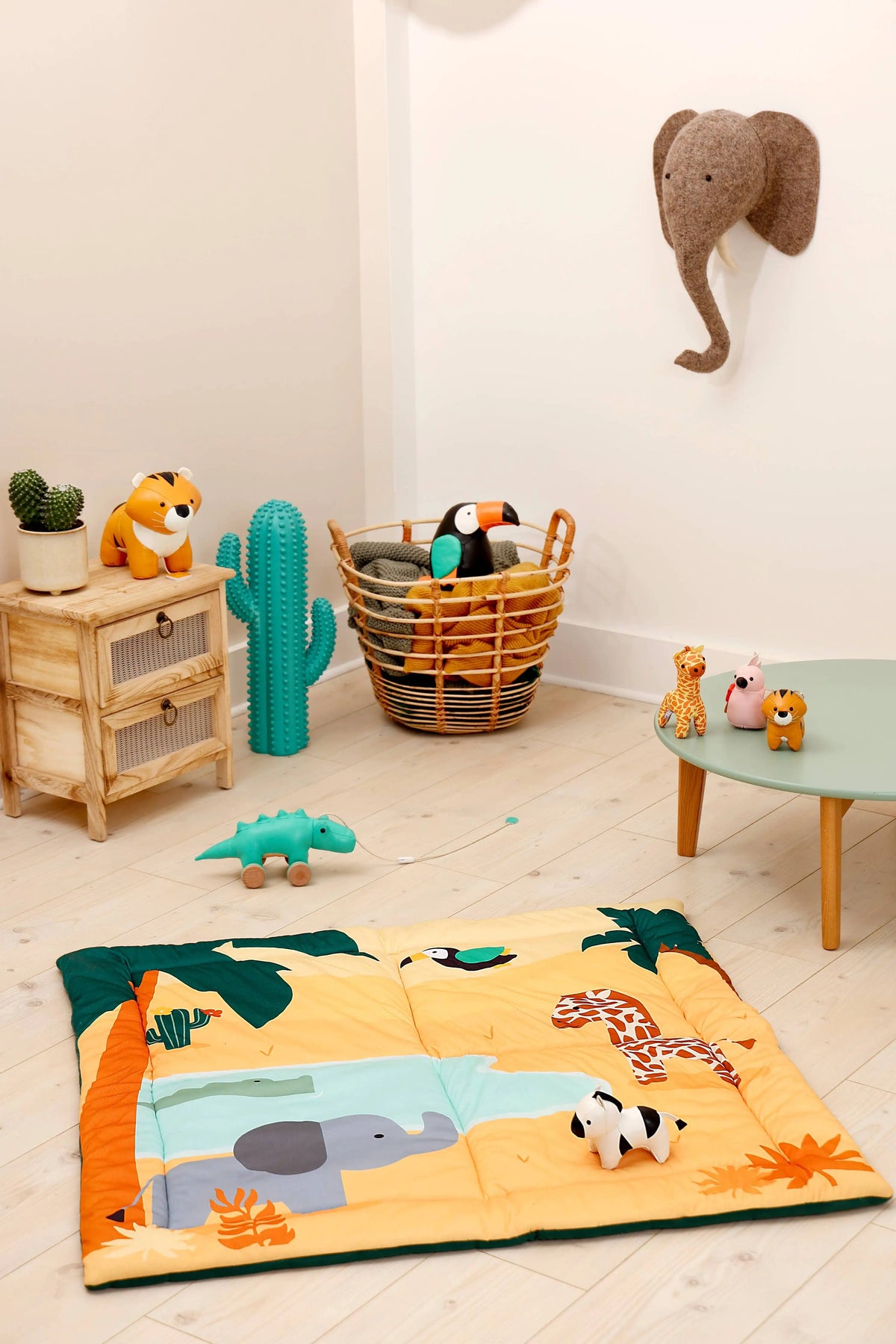 A colourful children's safari-themed floor mat with plush animal toys, displayed in a styled nursery room setting with a wooden side table, cactus, and wall-mounted elephant.