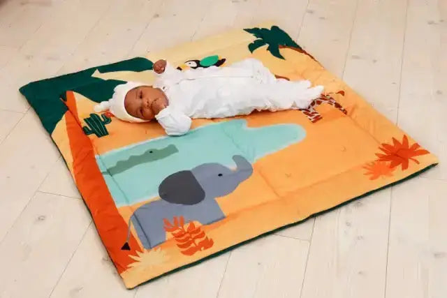 Baby lying on a colourful play mat with animal designs on a wooden floor.