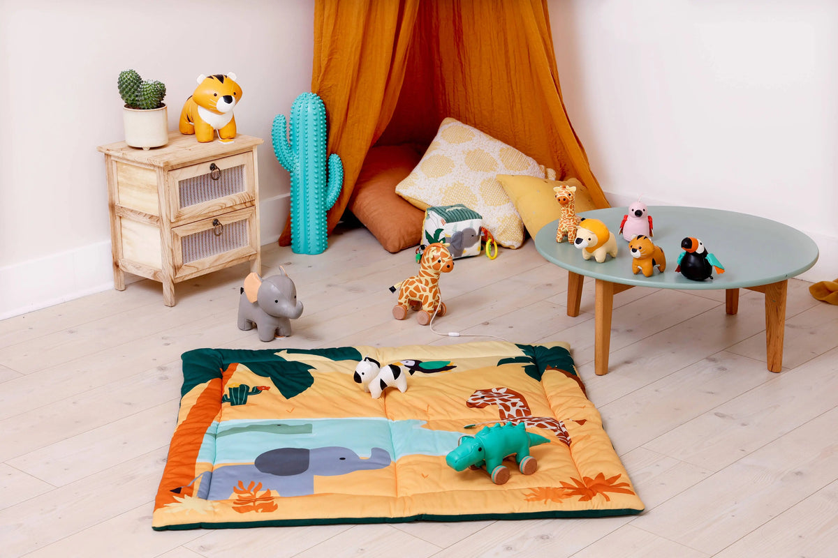 A colourful safari-themed play mat laid out on a light wood floor in a styled children's room. The play mat features illustrations of an elephant, crocodile, palm tree, and desert landscape, surrounded by plush safari animals including a giraffe, zebra, and elephant.