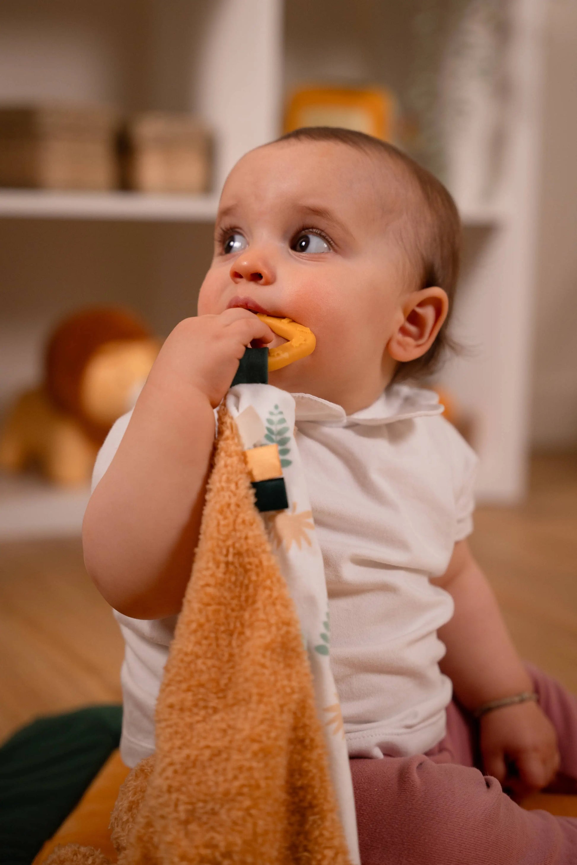 A baby wearing a white short-sleeve top and pink pants sits indoors while biting a yellow teething toy attached to a patterned cloth. The baby is holding a plush orange towel in the foreground, with a softly blurred room and shelves in the background.