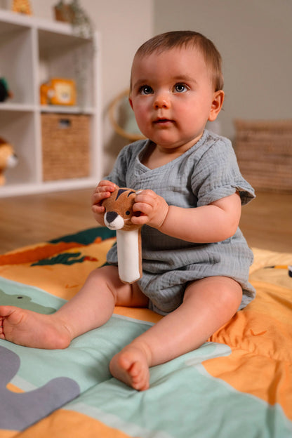Baby sitting on a colourful blanket holding a toy, with a shelf and toys in the background.