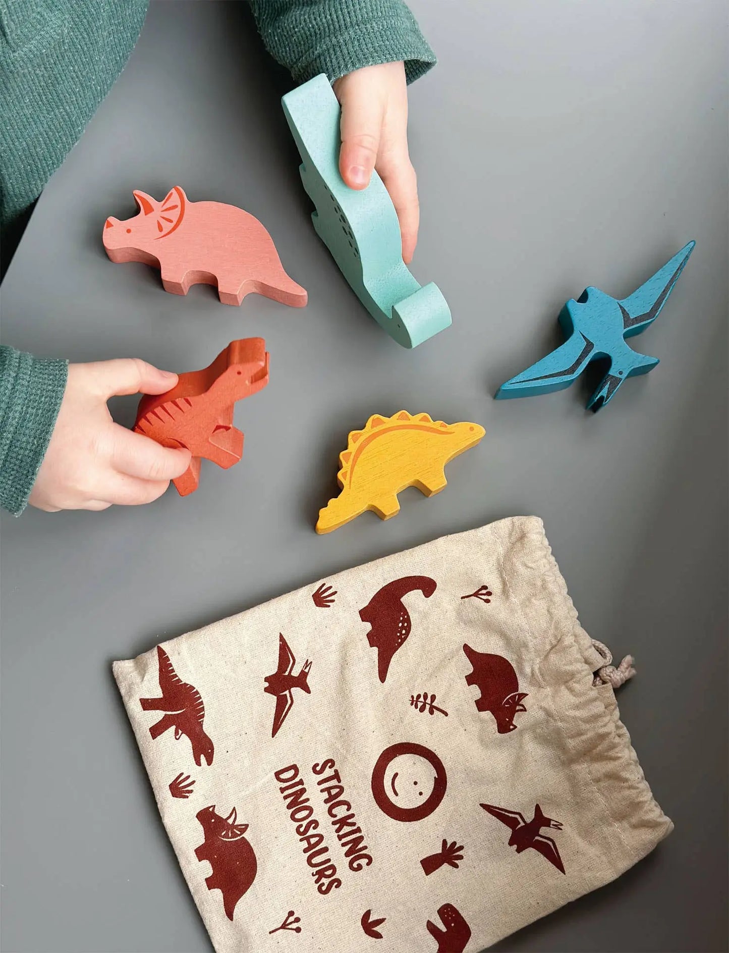 Children playing with colourful dinosaur-shaped toys on a grey surface, with a drawstring bag featuring dinosaur illustrations.