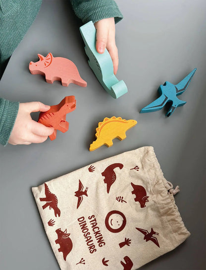 Children playing with colourful dinosaur-shaped toys on a grey surface, with a drawstring bag featuring dinosaur illustrations.