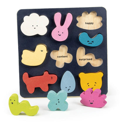 A wooden children's puzzle board with colorful cutout shapes and words printed on a white background. The board is dark blue with cutouts for a white cloud, pink bunny, tan cloud labeled 'happy', blue sad face, yellow chick, tan flower labeled 'content', tan cloud labeled 'surprised', red dog, light blue teardrop, yellow bear, and three loose matching pieces in green, light blue, and pink.