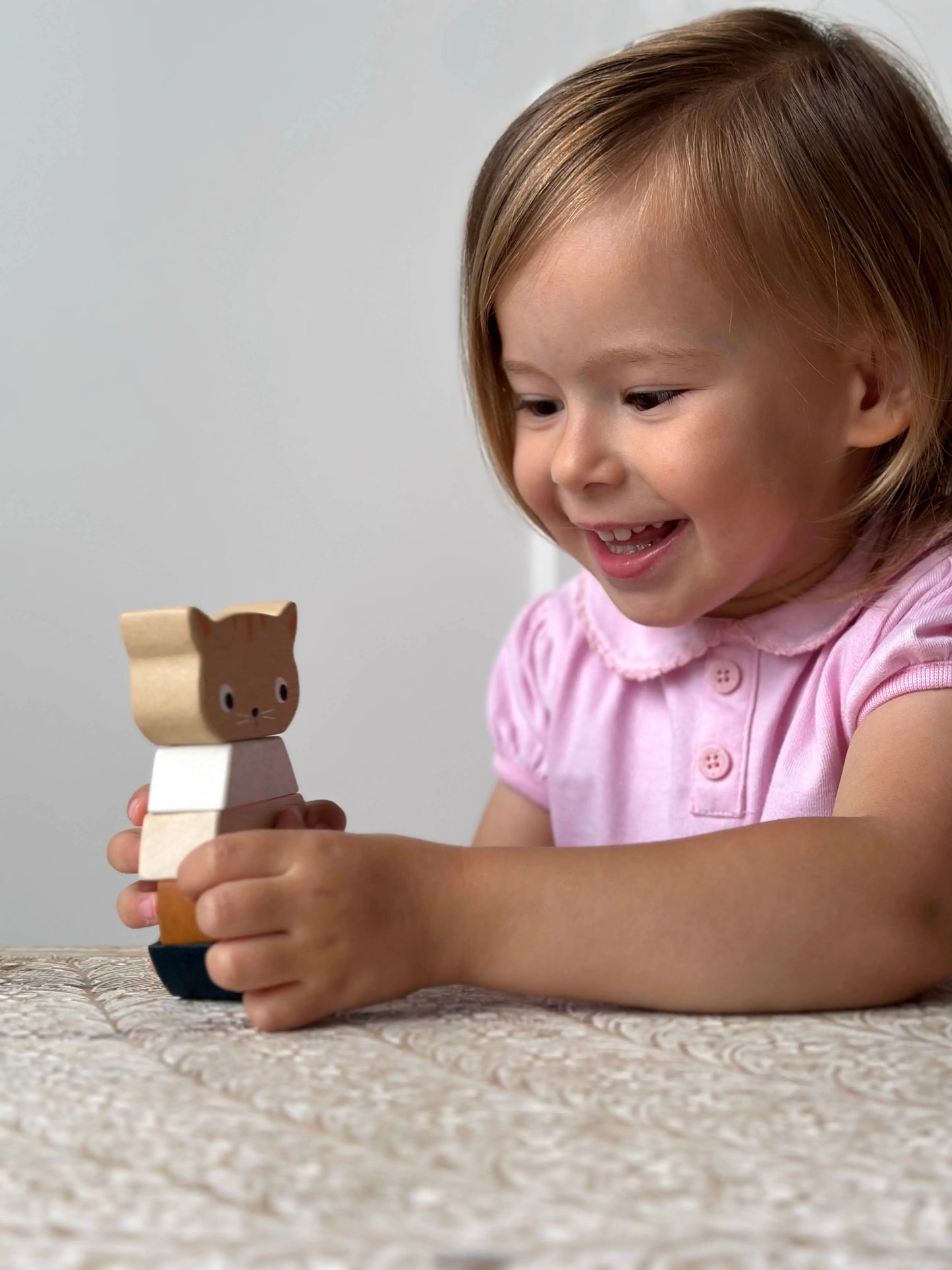 A young child with light brown hair, wearing a pink shirt, smiles while holding a small wooden animal toy with a brown cat head and white body on a textured cream surface.