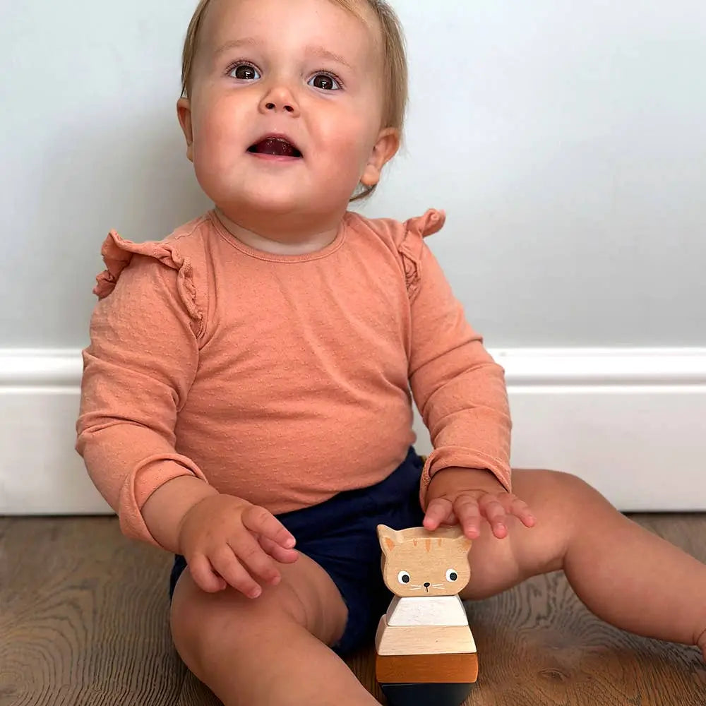 A baby sitting on a wooden floor against a white wall, wearing a peach long-sleeve top with ruffled shoulders and navy shorts, holding a small wooden kitten toy.
