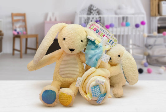 A plush baby bunny toy with long ears and blue/yellow feet, shown with a rolled fabric accessory and a hanging tag in a softly blurred nursery-like setting.